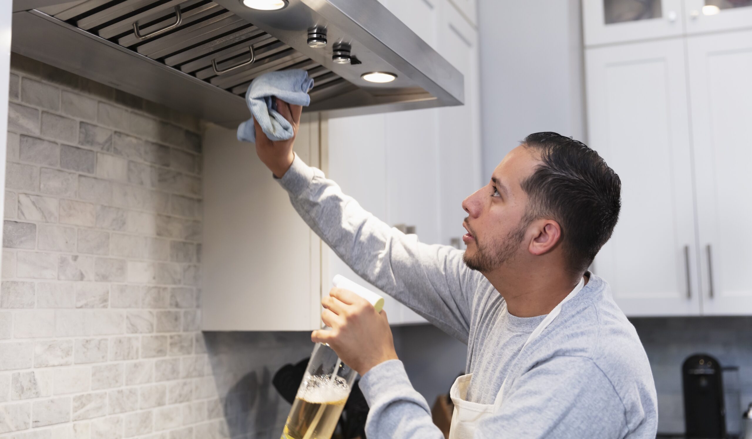 An Hispanic man in his 30s doing housework. He is standing in the kitchen, cleaning the range hood. He could be the homeowner doing chores, or a cleaner from a housekeeping service.